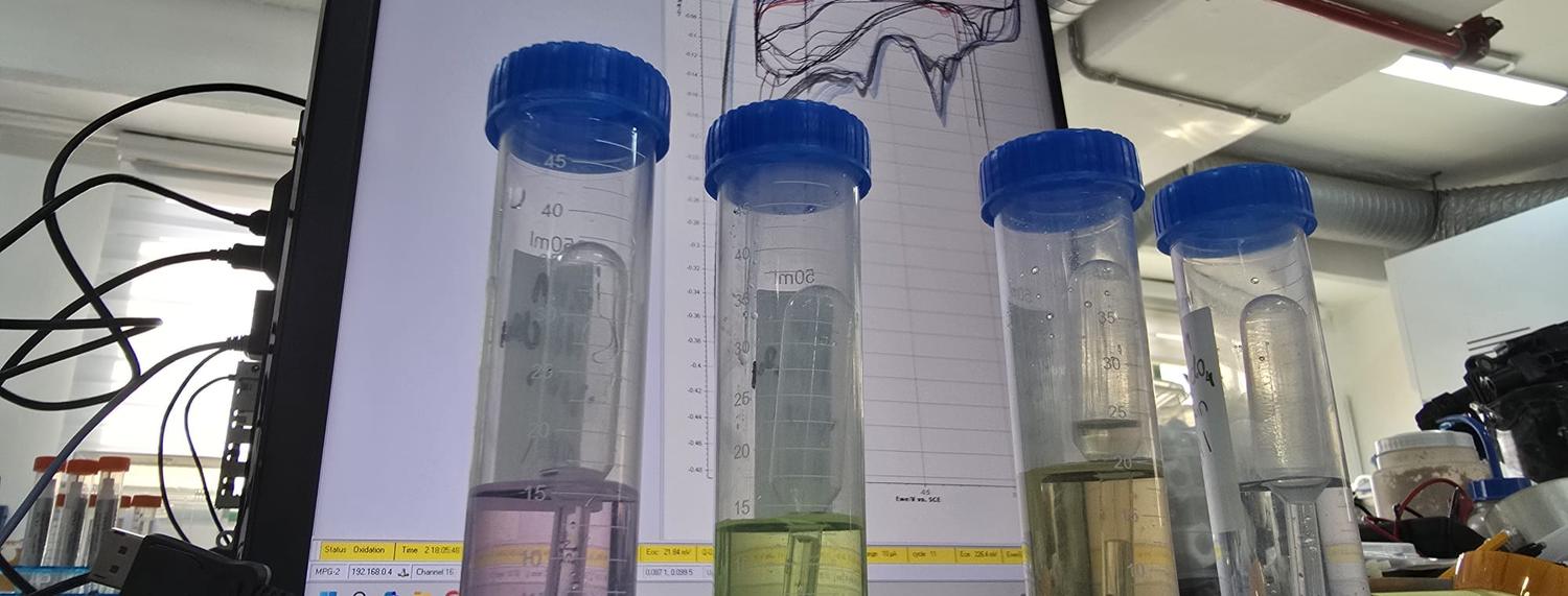 Four rare earth sample solutions in graduated vials in front of a monitor showing cyclic voltammetry traces